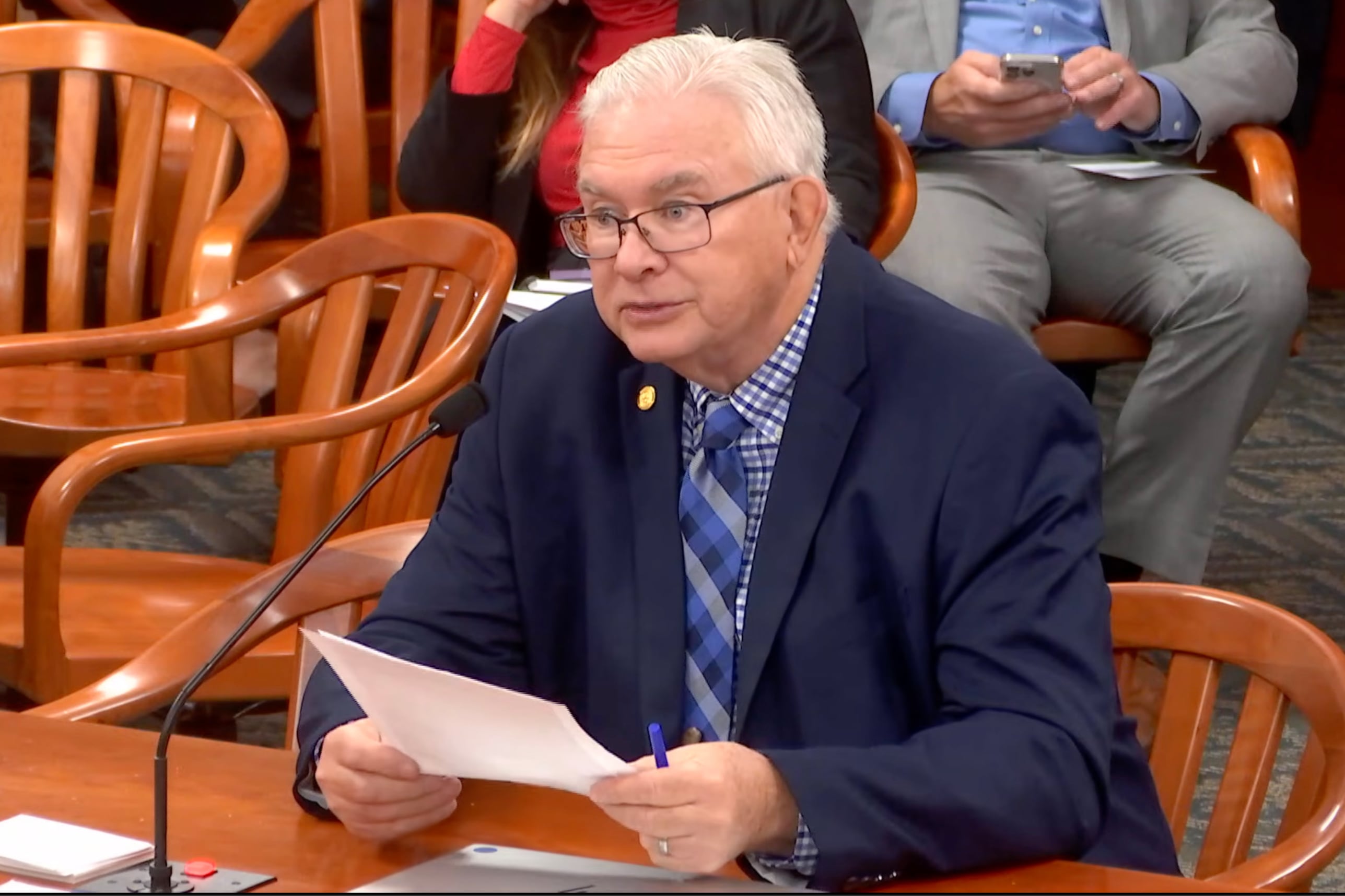 A screen grab of a white man in a dark suit sitting at a wooden table reading from a piece of paper into a microphone with two people sitting in chairs in the background.