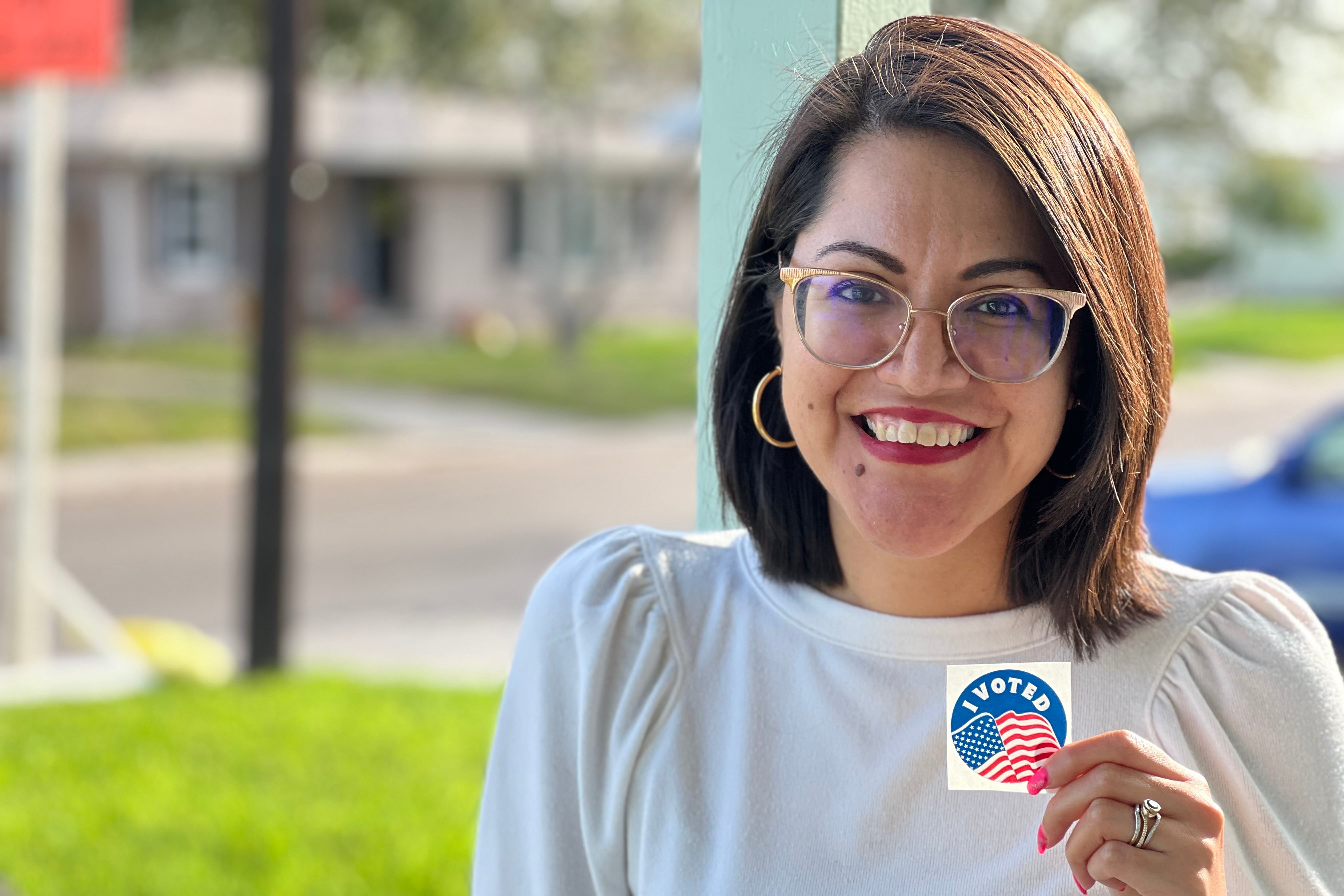 A woman with short brown hair and wearing a white blouse holds a sticker that says "I Voted" while posing for a photograph outside.