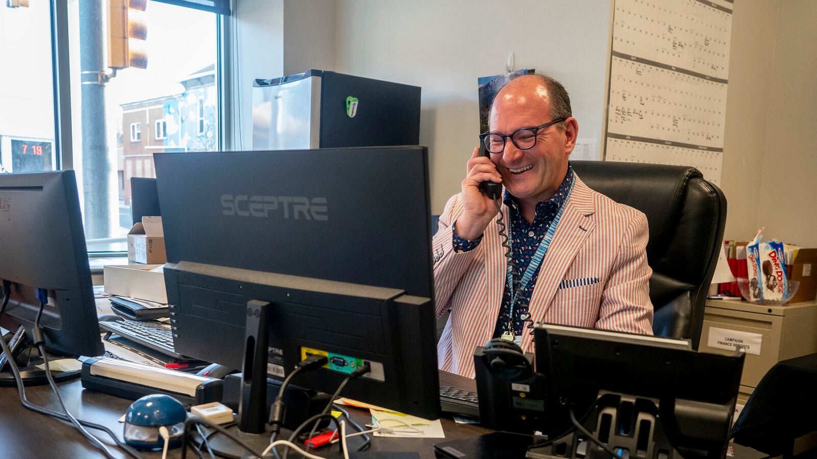 a man wearing a red and white striped suit answers the phone while sitting at a desk behind a computer screen.