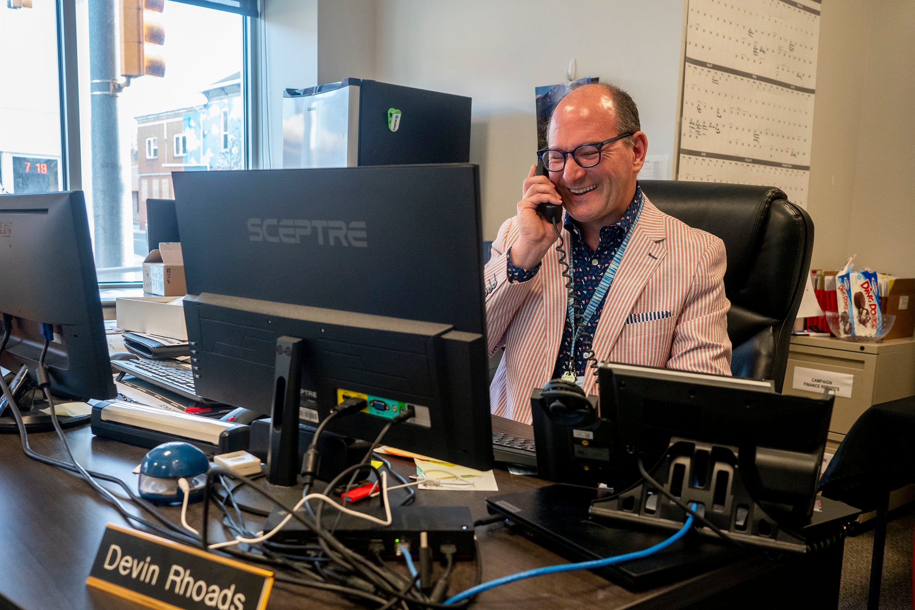 a man wearing a red and white striped suit answers the phone while sitting at a desk behind a computer screen.