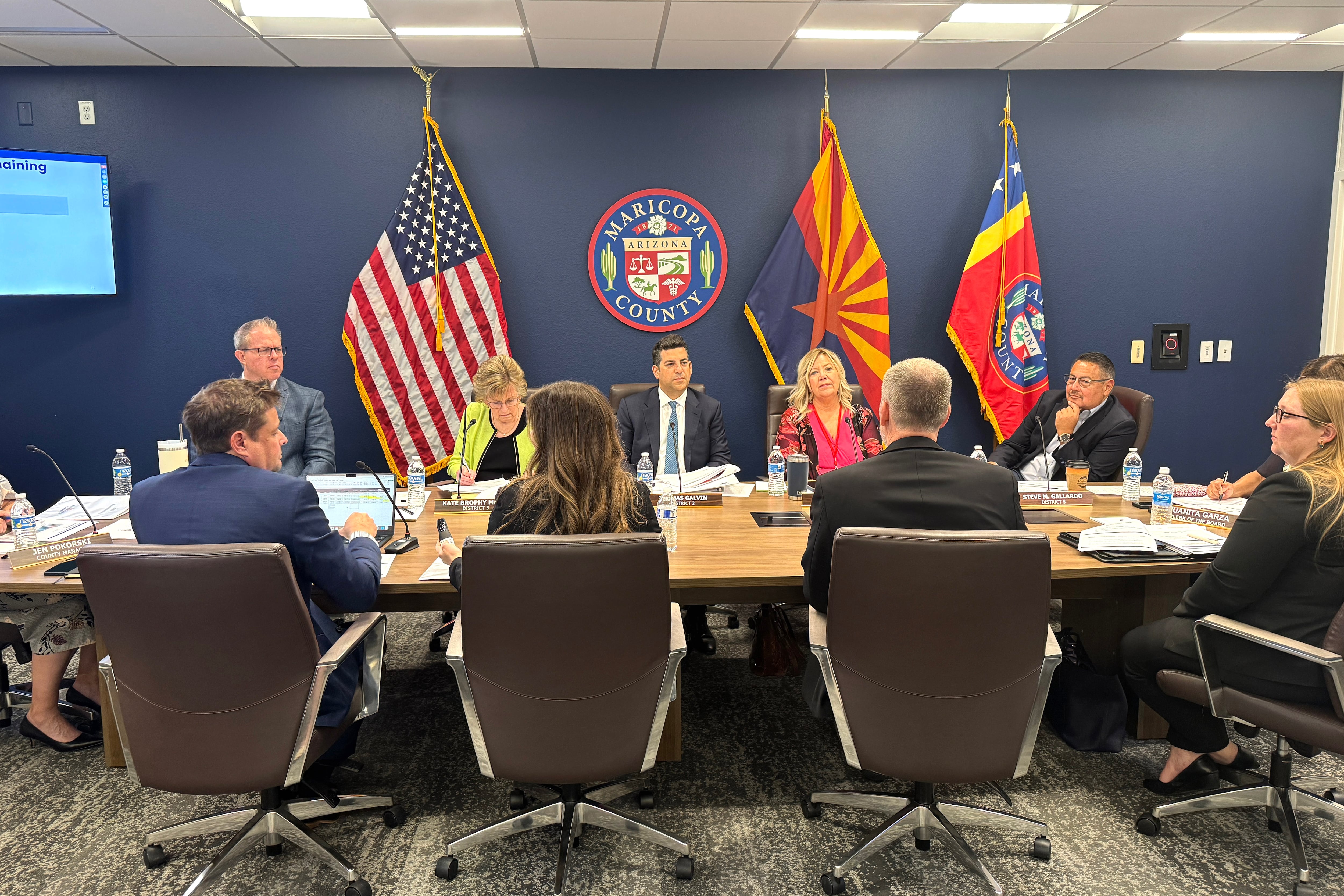 People in business attire sit around a table with flags in the background.