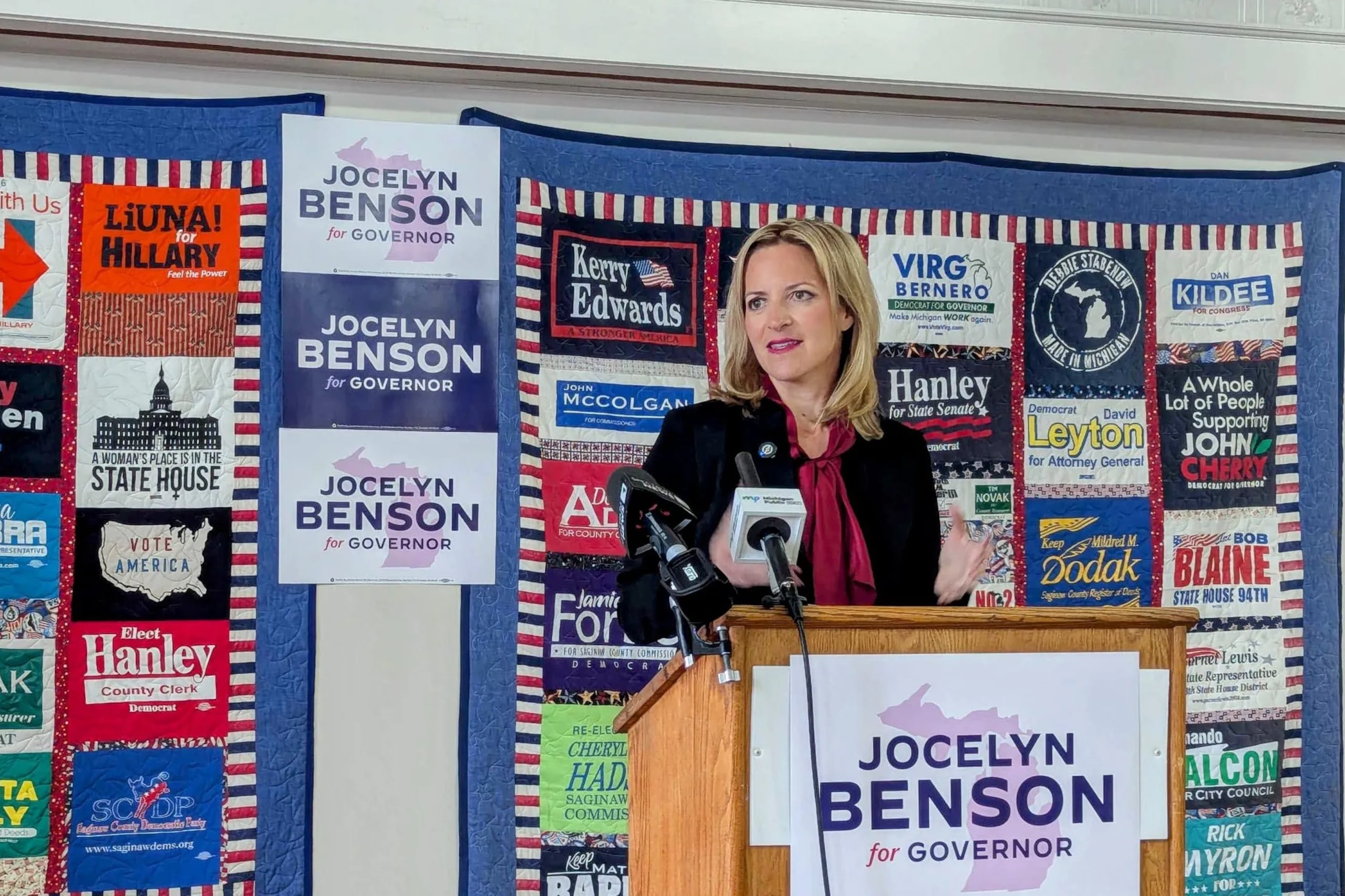 A photograph of a white woman in a suit standing behind a wooden podium and in front of a large wall covered in campaign posters.