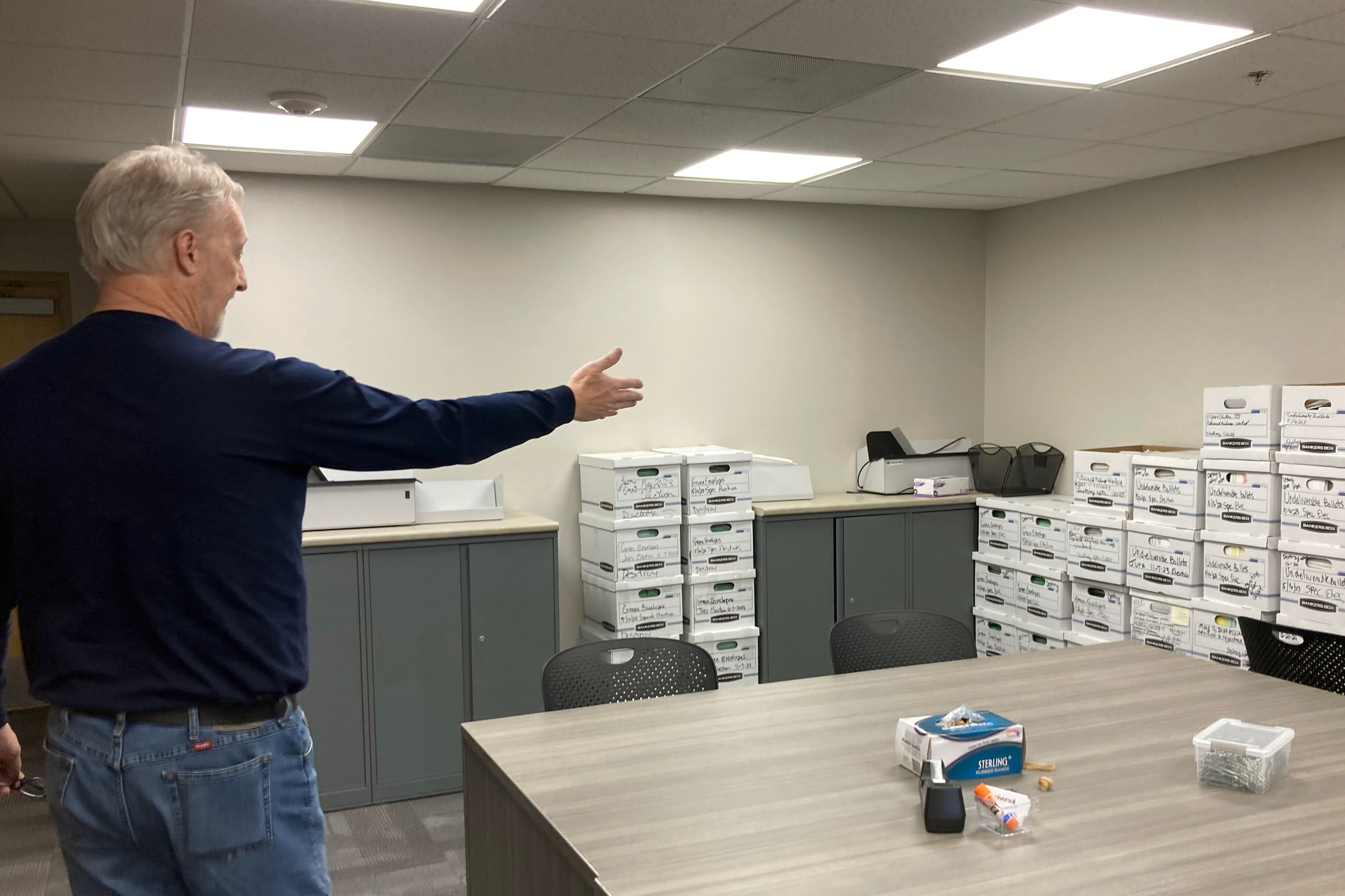 A man wearing a dark sweater points to stacks of white boxes in a room with tan walls and a giant wooden desk in the foreground.