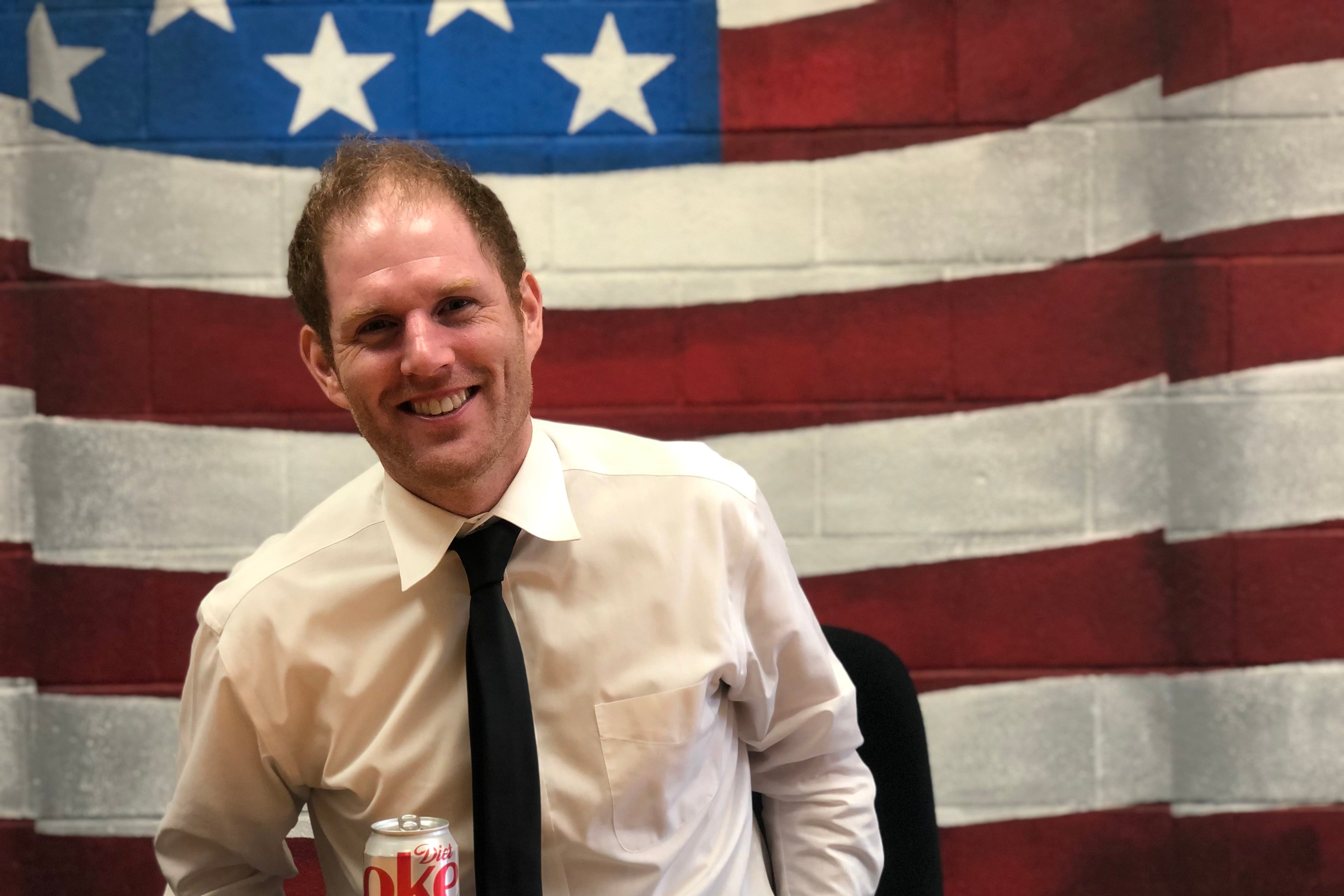 A man in a shirt and tie sits at a desk with an American flag painted on the wall behind him