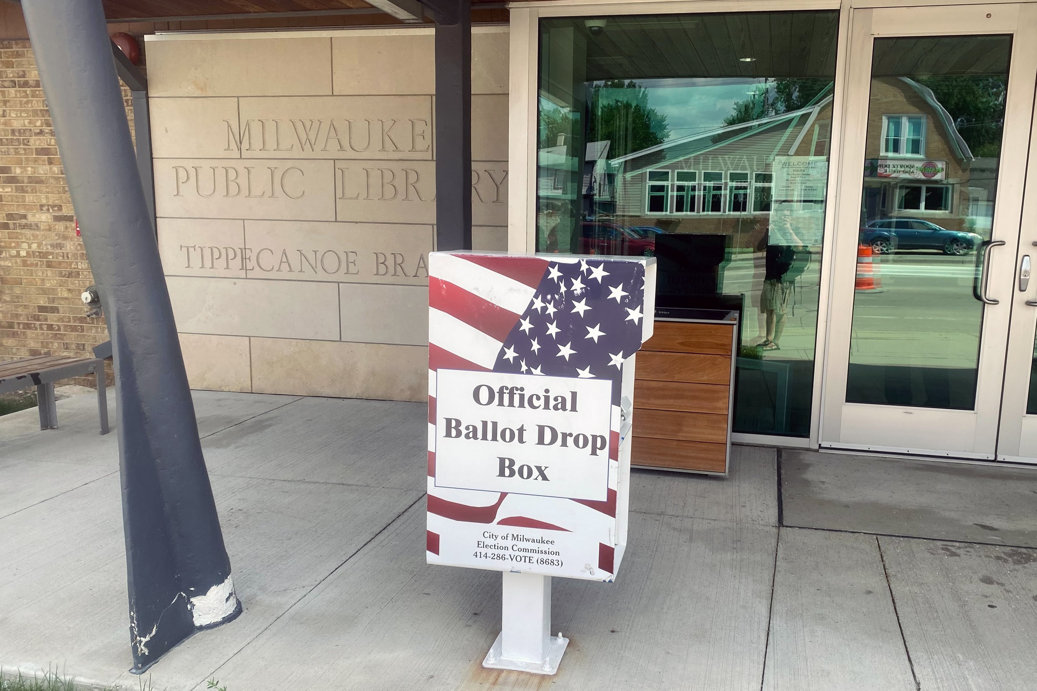 A metal box covered in the American flag stands outside of a stone library.