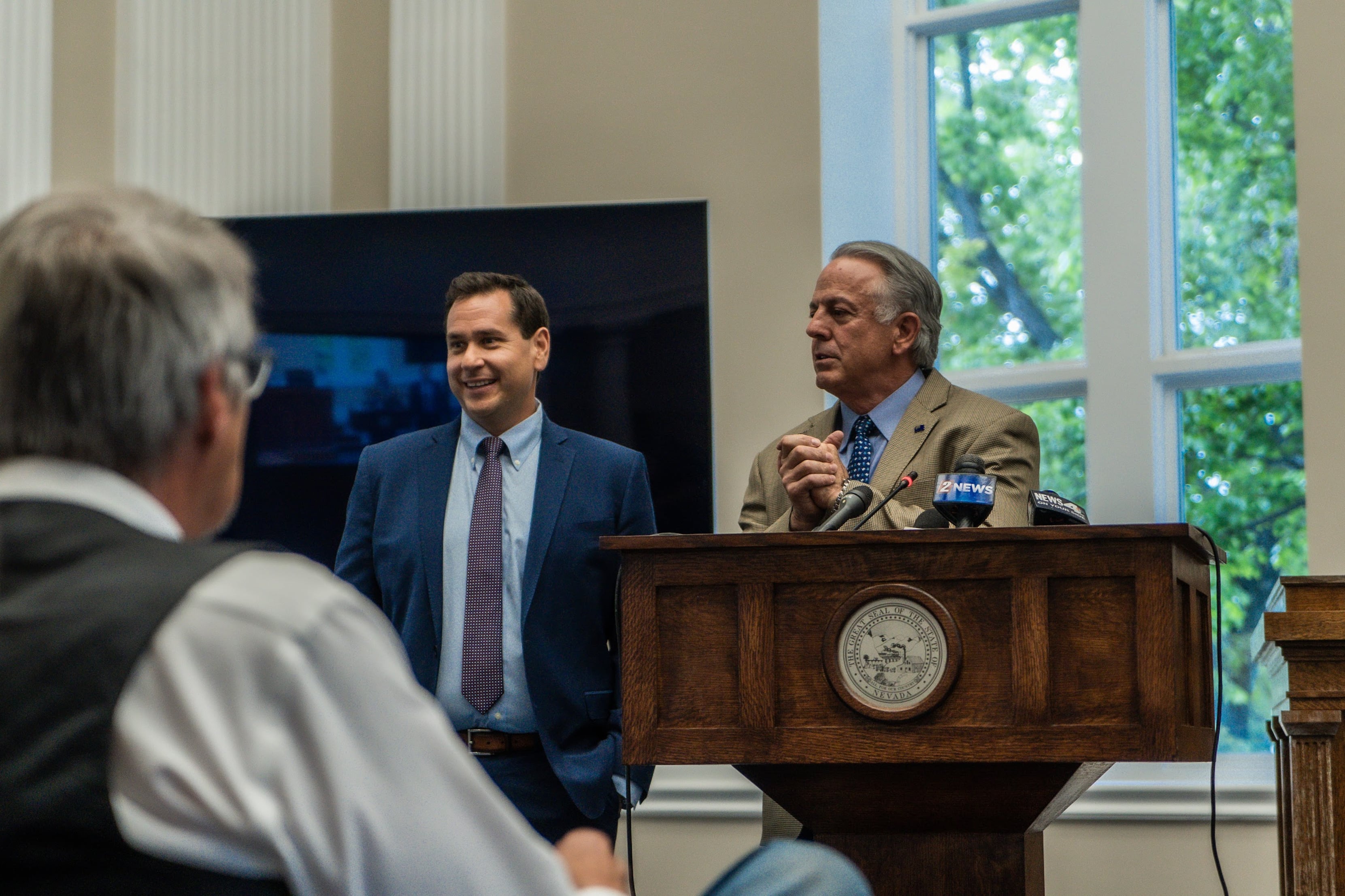 A man in a blue suit and tie stands next to another man in a beige jacket and tie who is behind a podium. 