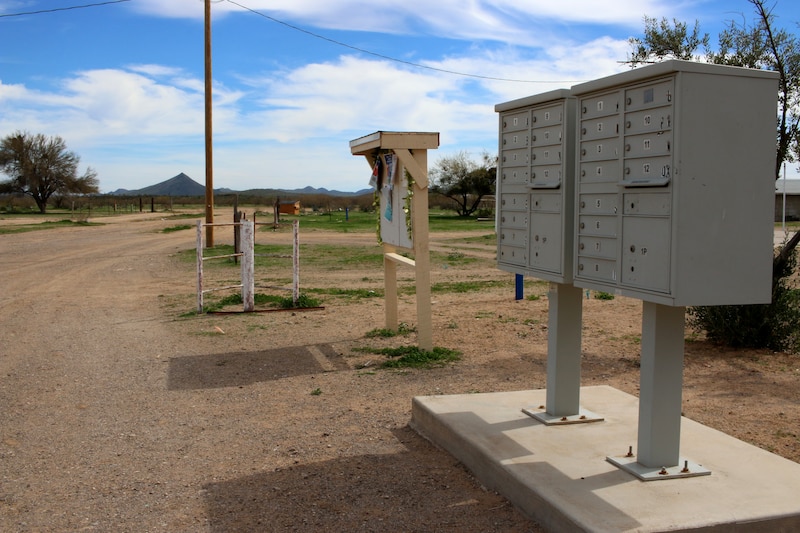 A cluster of mailboxes stand near a dirt road with a wooden sign holder in the middle ground with a mountain and blue sky in the background.