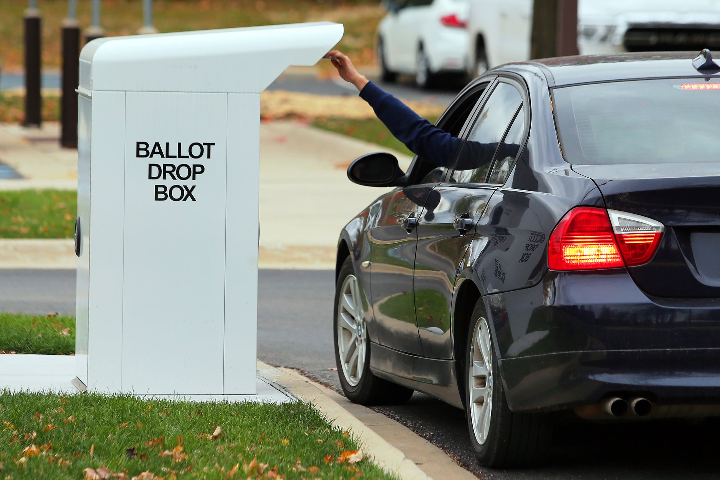 A white box with the words "Ballot Drop Box" on the side with an arm reaching out from a car window to drop off a ballot.