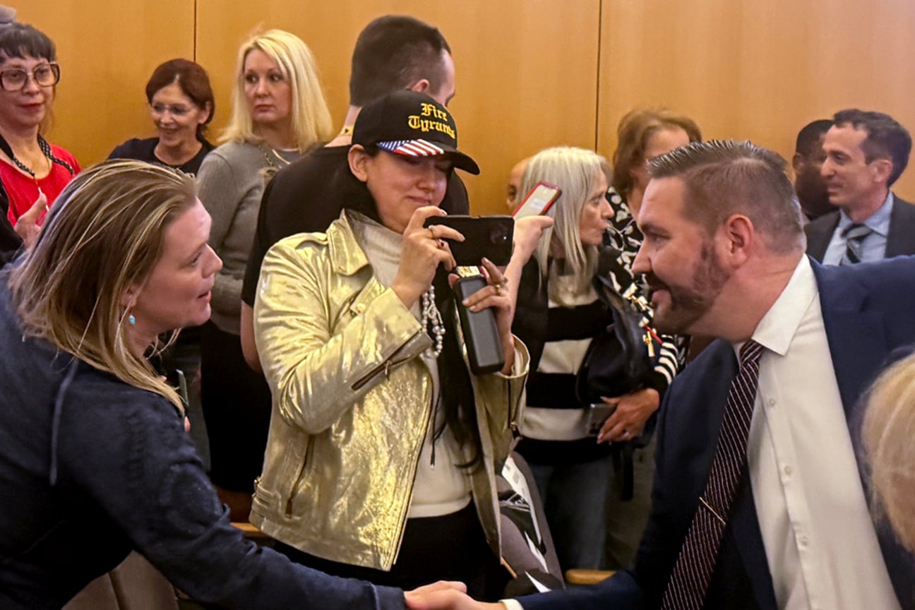 A man in a dark suit jacket reaches to shake hands with someone in a dark jacket.