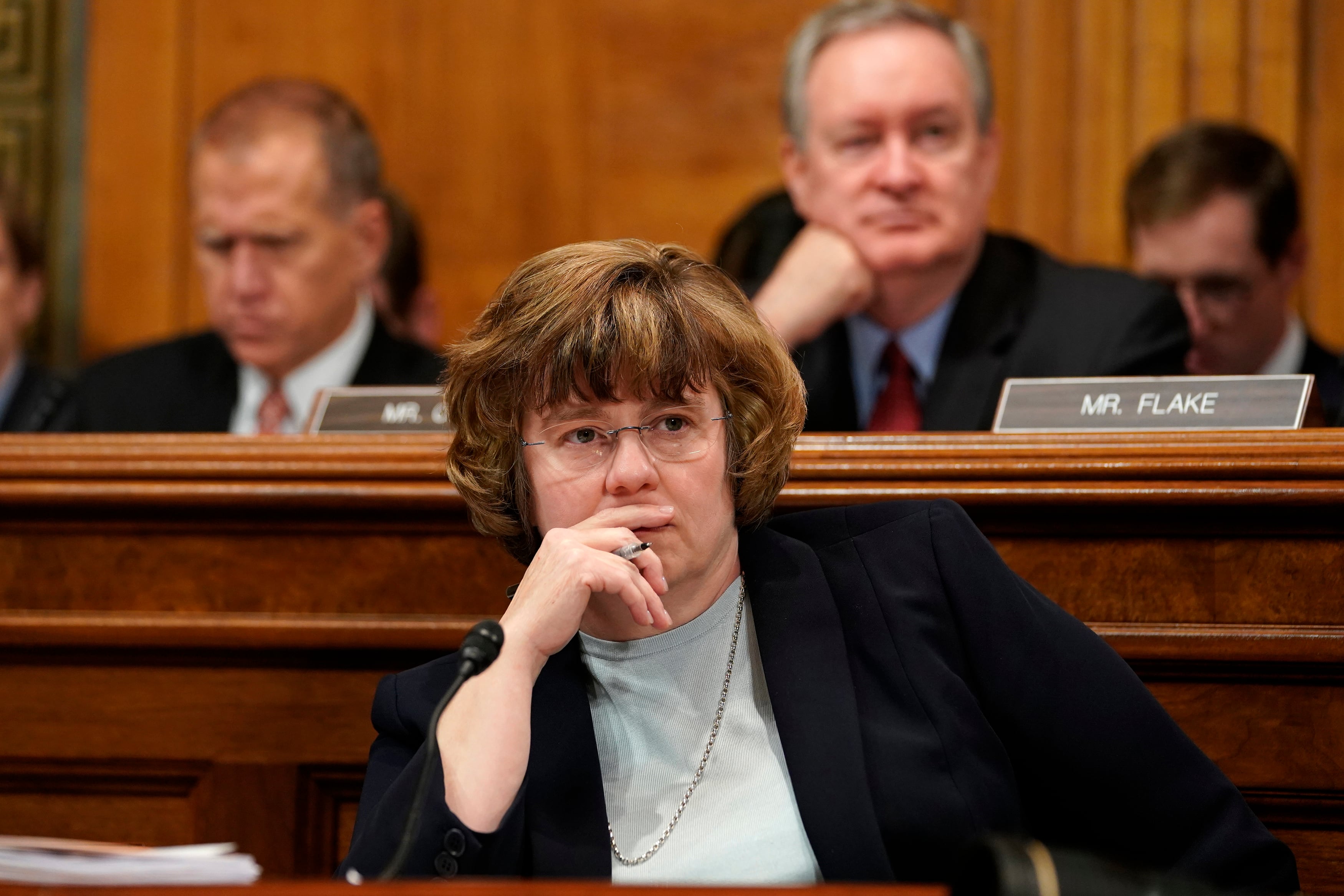 A white woman with short hair and wearing a suit jacket rests her chin on her hand while listening in a large room.