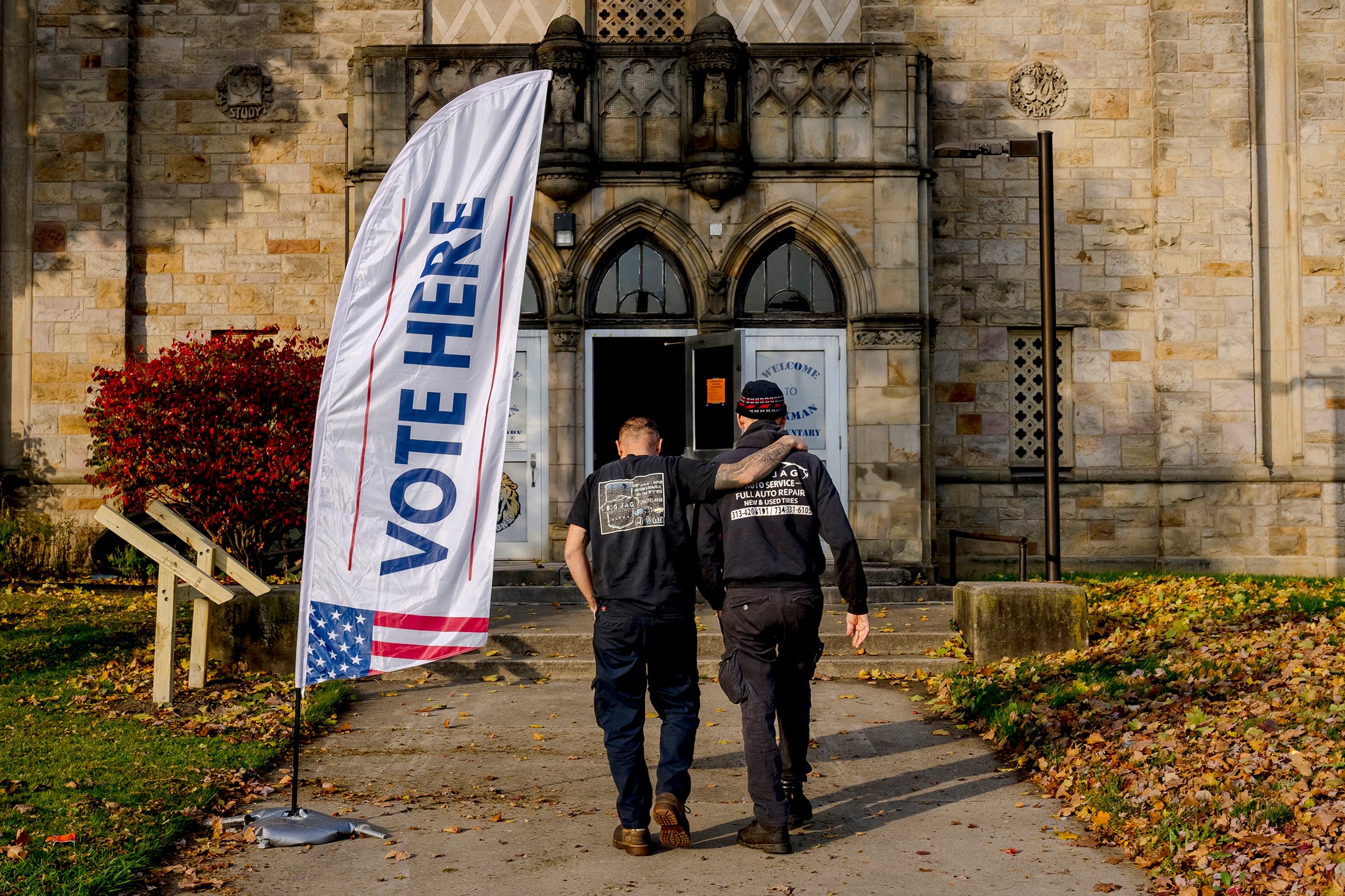 Two people walk in tandem into the front entrance of a large stone building. There is a long white flag that reads "VOTE HERE," next to the two people.