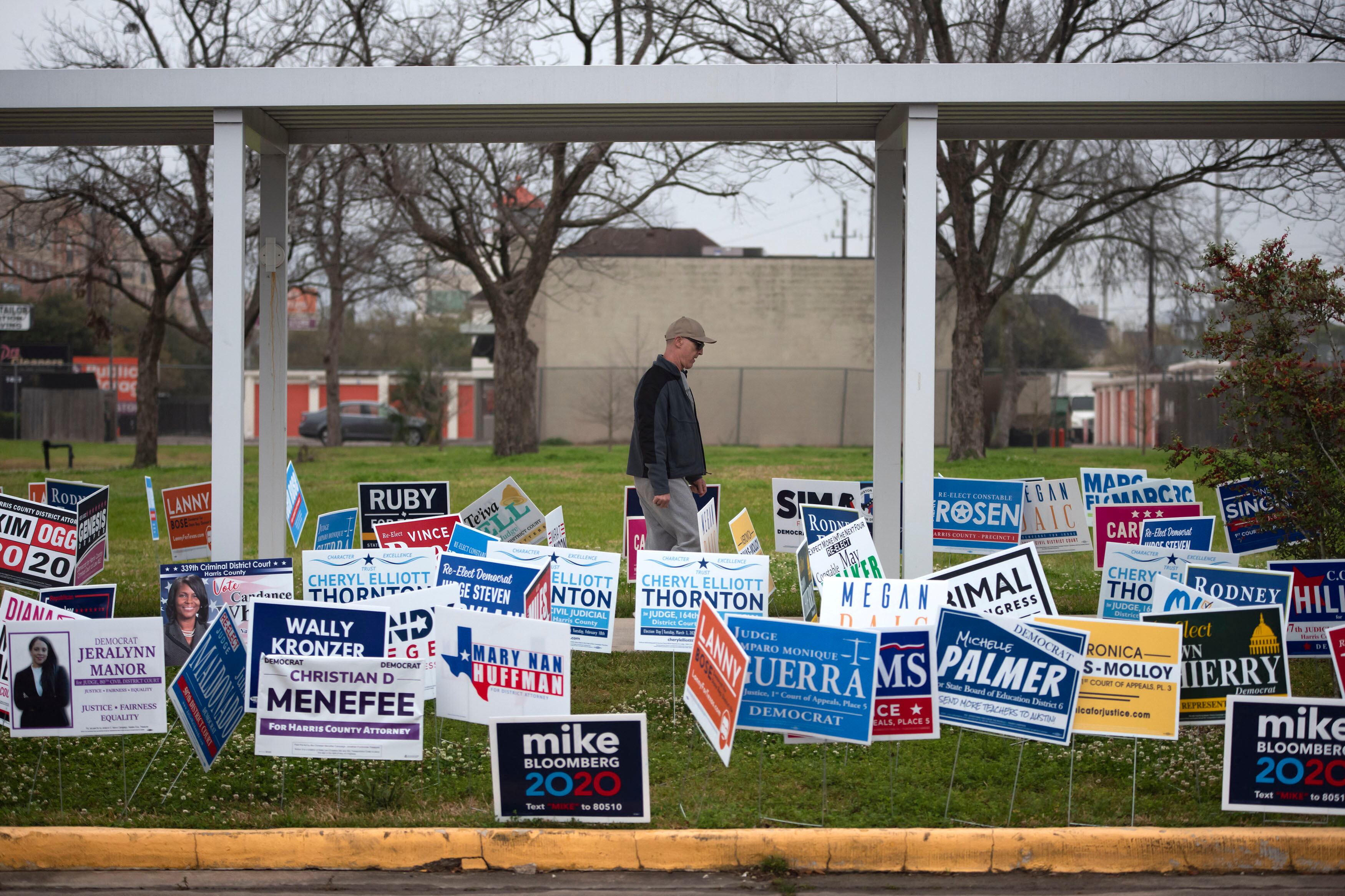 A person wearing a hat and a dark jacket walks behind a lawn full of candidate signs with a white structure over the top and a building in the background.
