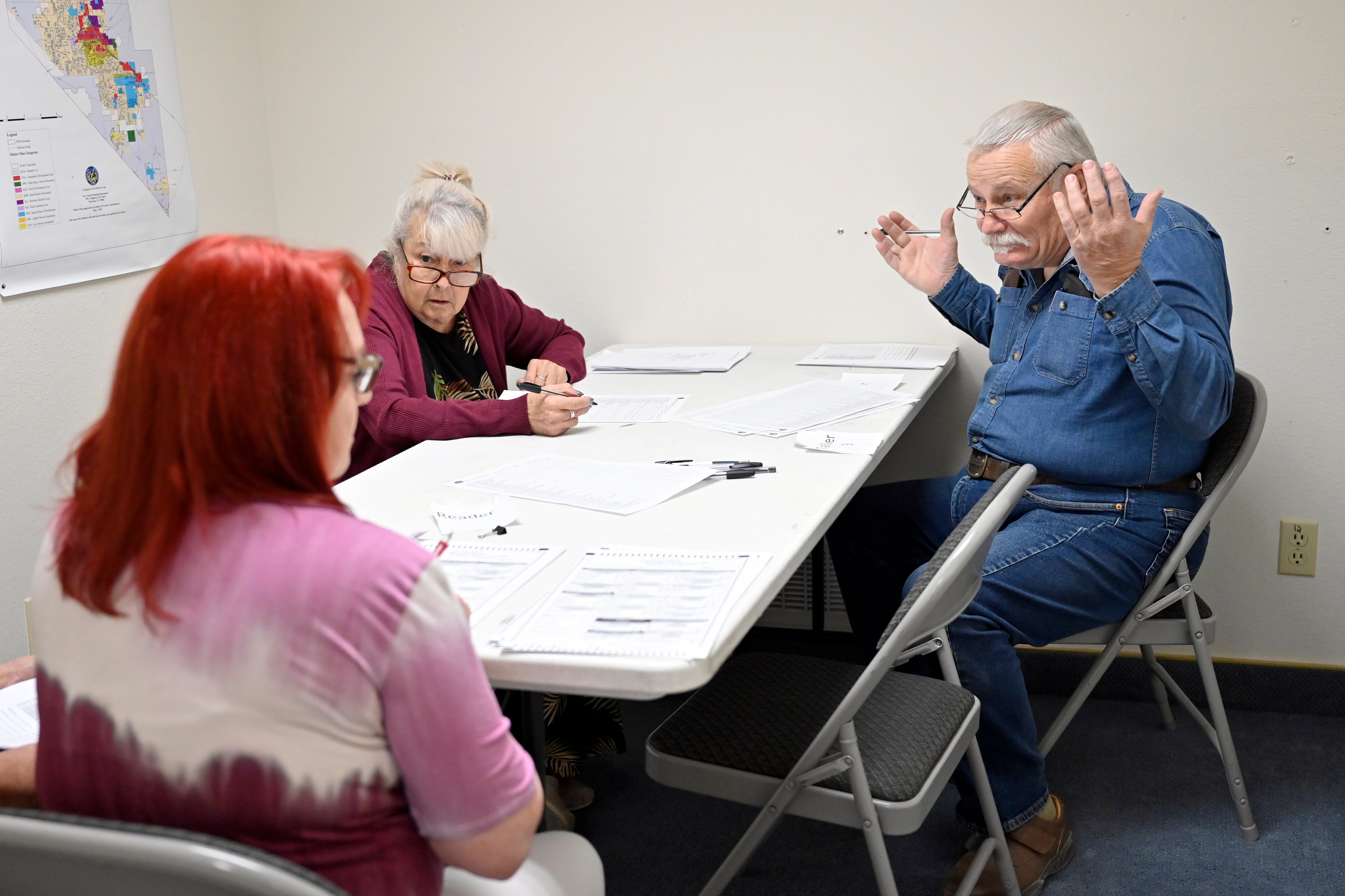 Three people sit around a white table surrounded by folding chairs. A man wearing a denim shirt throws his hands up.