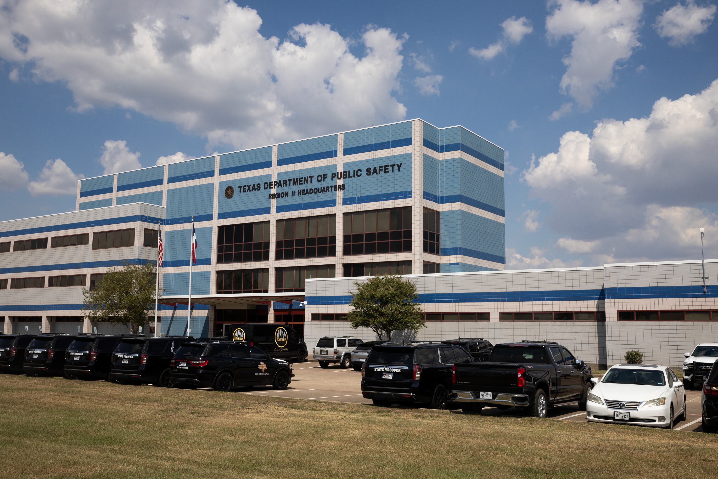 A photograph of a large blue and white building with a parking lot of cars with clouds in a blue sky.