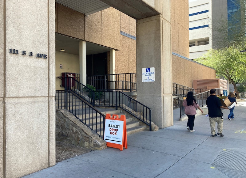 People walk by the entrance to a large stone building with an orange and white sign sitting out front that reads "Ballot Drop Box".