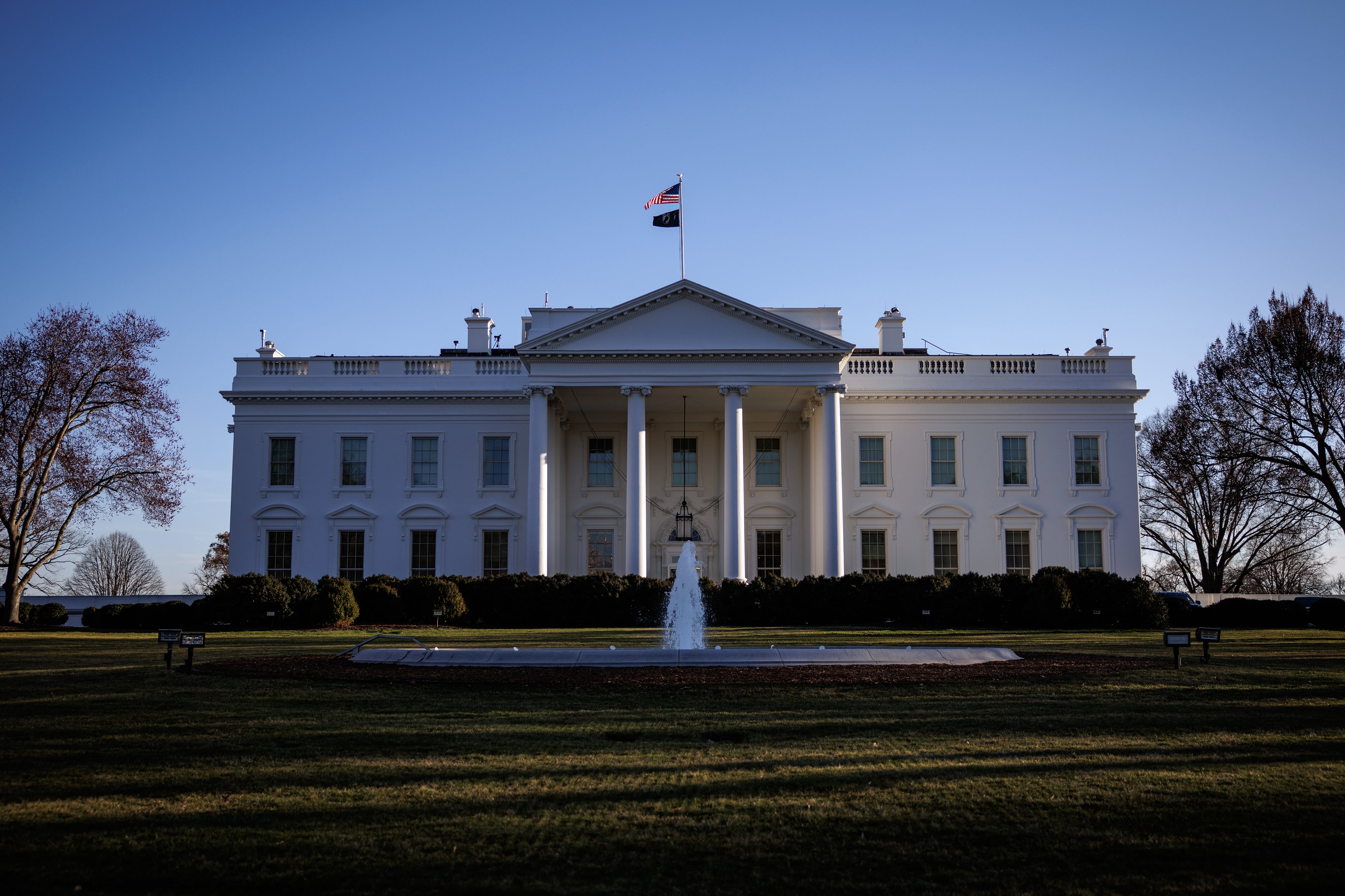 A front view of the White House with trees on each side of the building and a water fountain and a grassy lawn in the foreground.