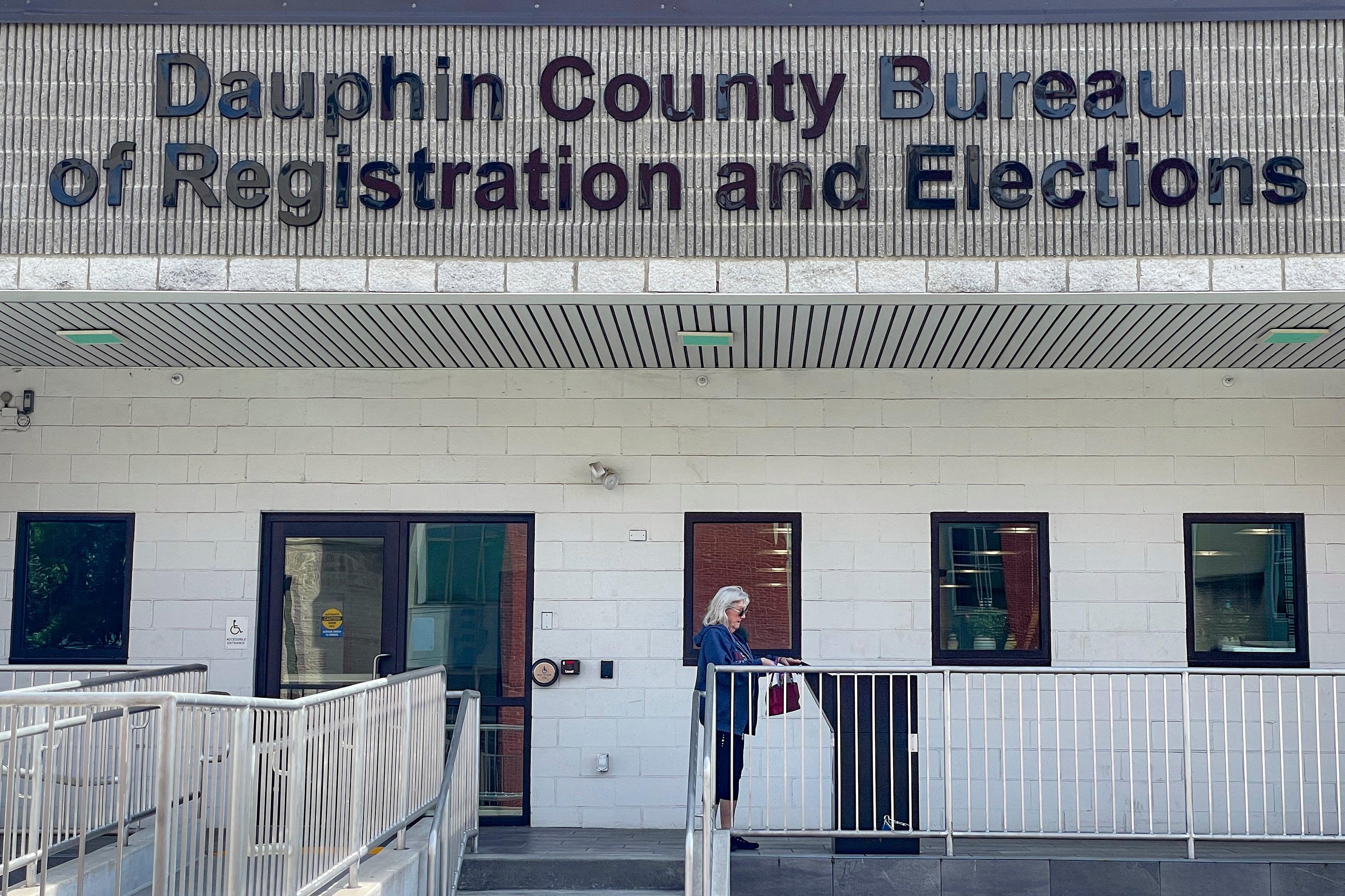 A person with short white hair stands next to a ballot drop box in front of a building with the words "Dauphin County Bureau of Registration and Elections."