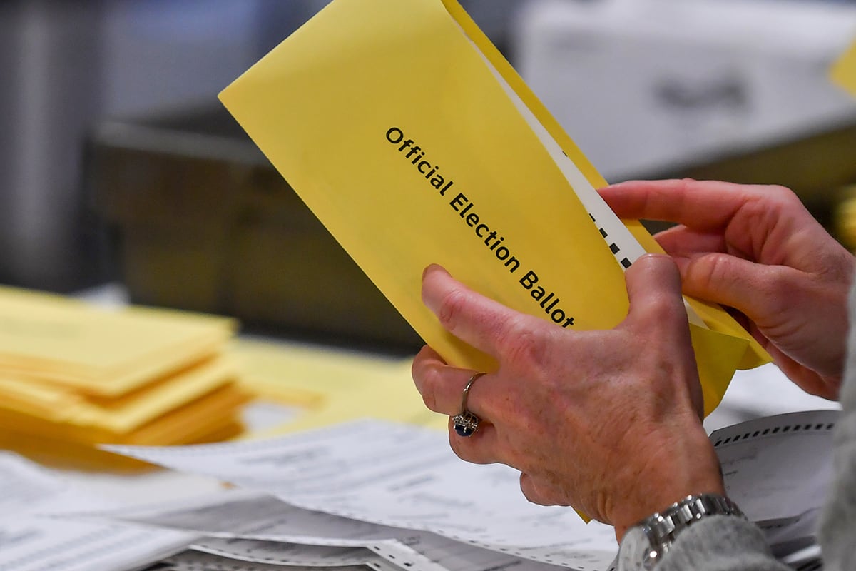 A pair of hands wearing rings holds a yellow envelope with the words " Official Election Ballot," with other ballots and papers in the background.