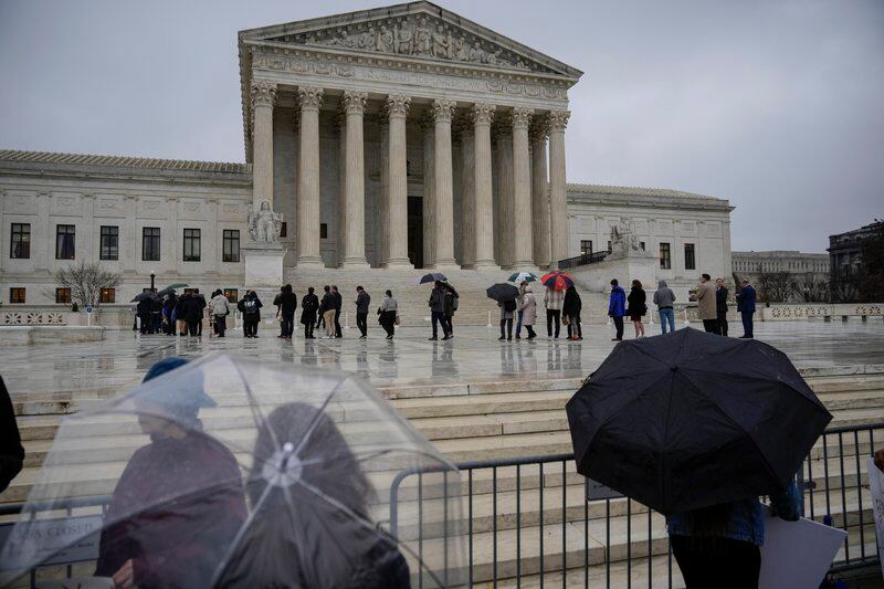 People stand in line, some holding umbrellas, across the plaza in front of the U.S. Supreme Court.